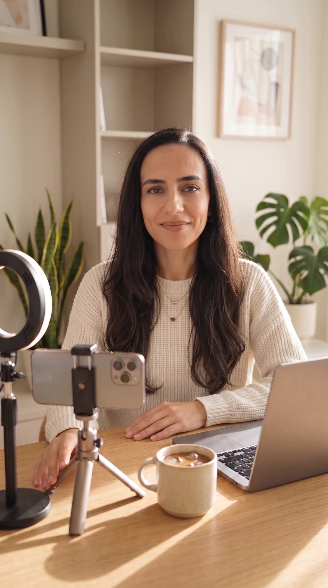 Seda Akyurek in a cream sweater at a sunlit desk with her laptop, an iced coffee and a phone on a tripod